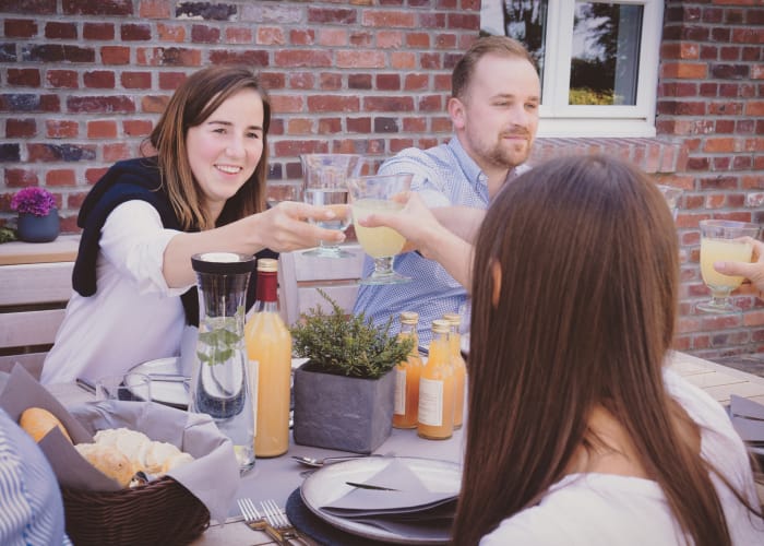 Junge Menschen sitzen lachend auf einer Terrasse und stoßen mit Getränken an. Die Stimmung ist gut und der Tisch ist reichlich gedeckt. Zu sehen sind unter anderem Brot, Apfelsaft, Wasser und Geschirr.
