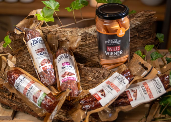 Diverse Produkte von der Marke „Wilder Heinrich“, welche Wildprodukte herstellt und vertreibt. Pfefferbeißer, Salami und Wiener werden auf einem Holzbouquet mit Efeu präsentiert.